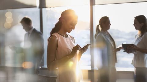 Woman looking down at her phone in an office setting with people talking behind her.